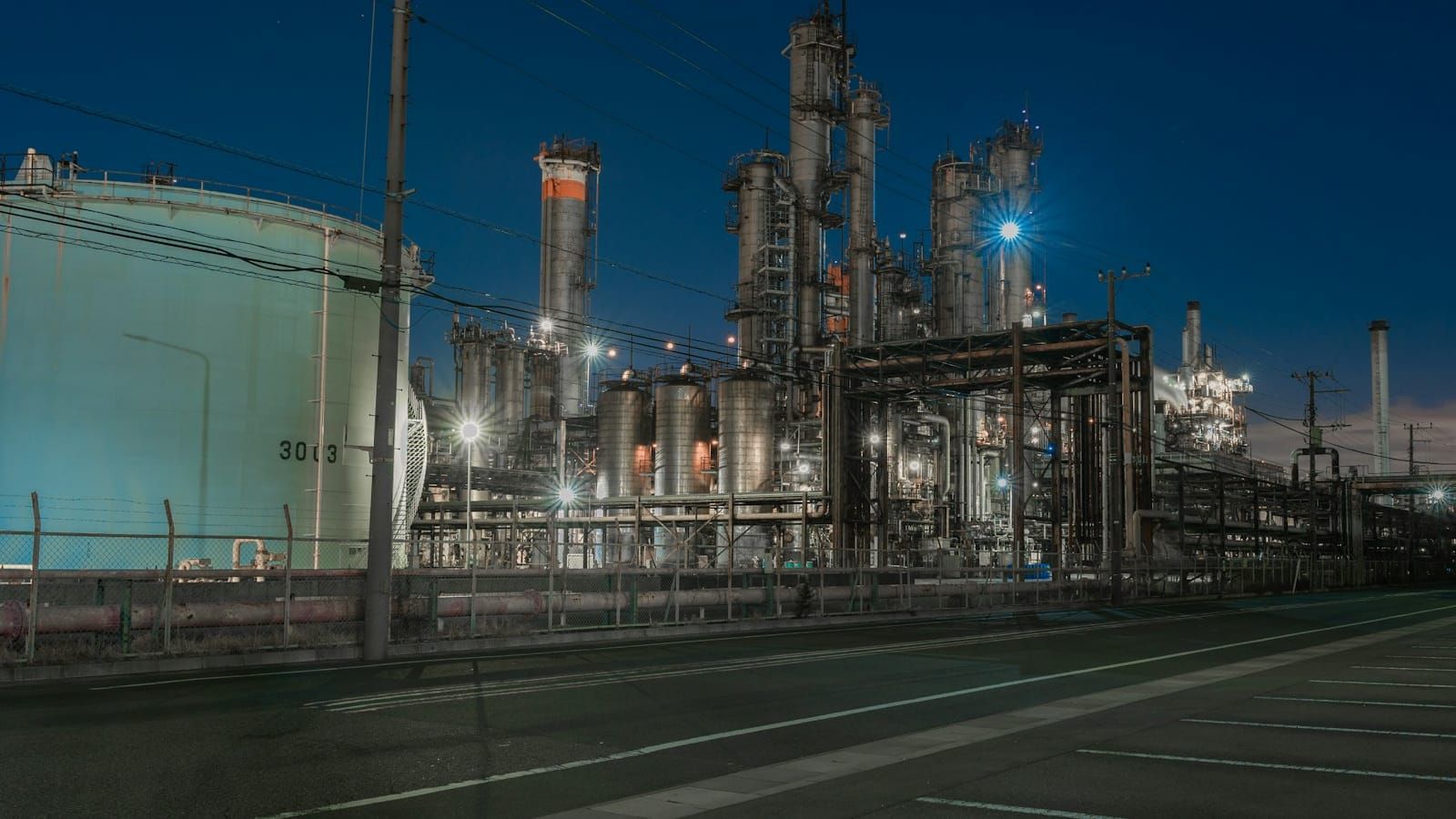 A refinery at blue hour: dense pipes, towers and tanks lit up against the night sky