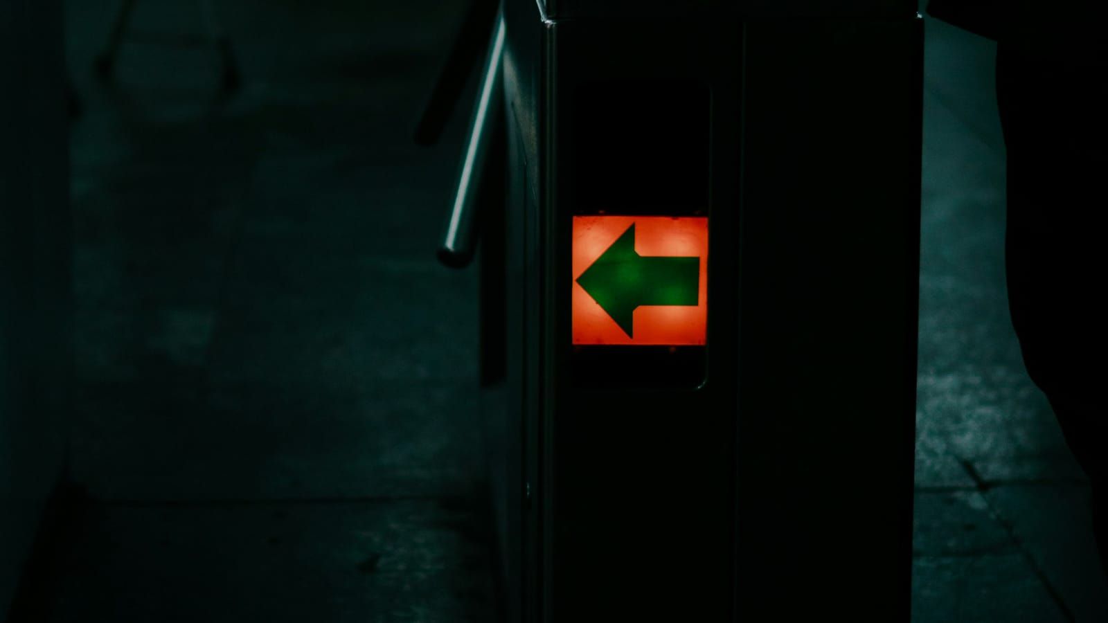 A turnstile in a dim station, its side lit by a red sign with a green arrow