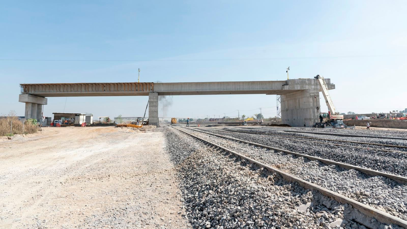 A railway construction site with new tracks laid on gravel ballast, a half-built concrete overpass and a crane in the distance, and no train on the line yet