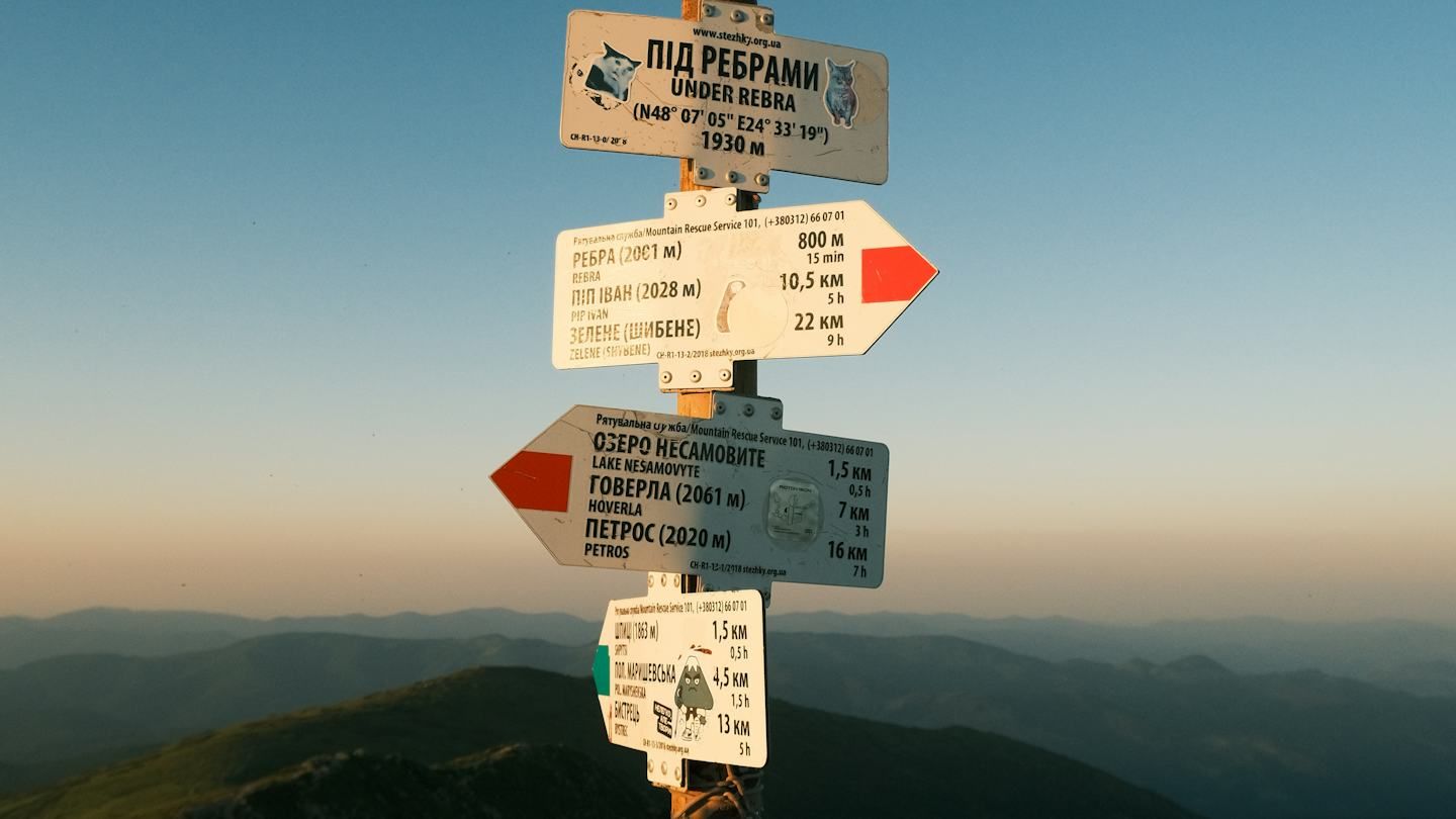 A trail signpost on a mountain top at dusk, its white boards pointing left and right with distances and walking times to each destination, ridges fading into the distance