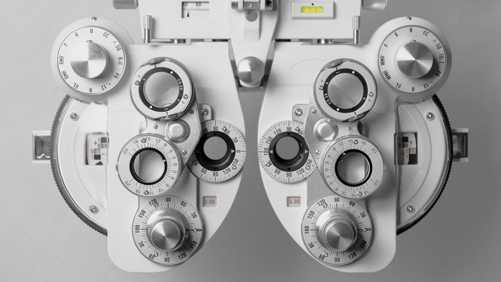 Front view of a white phoropter, two mirrored banks of lens dials, each opening ringed by scales and metal knobs, against a light grey background