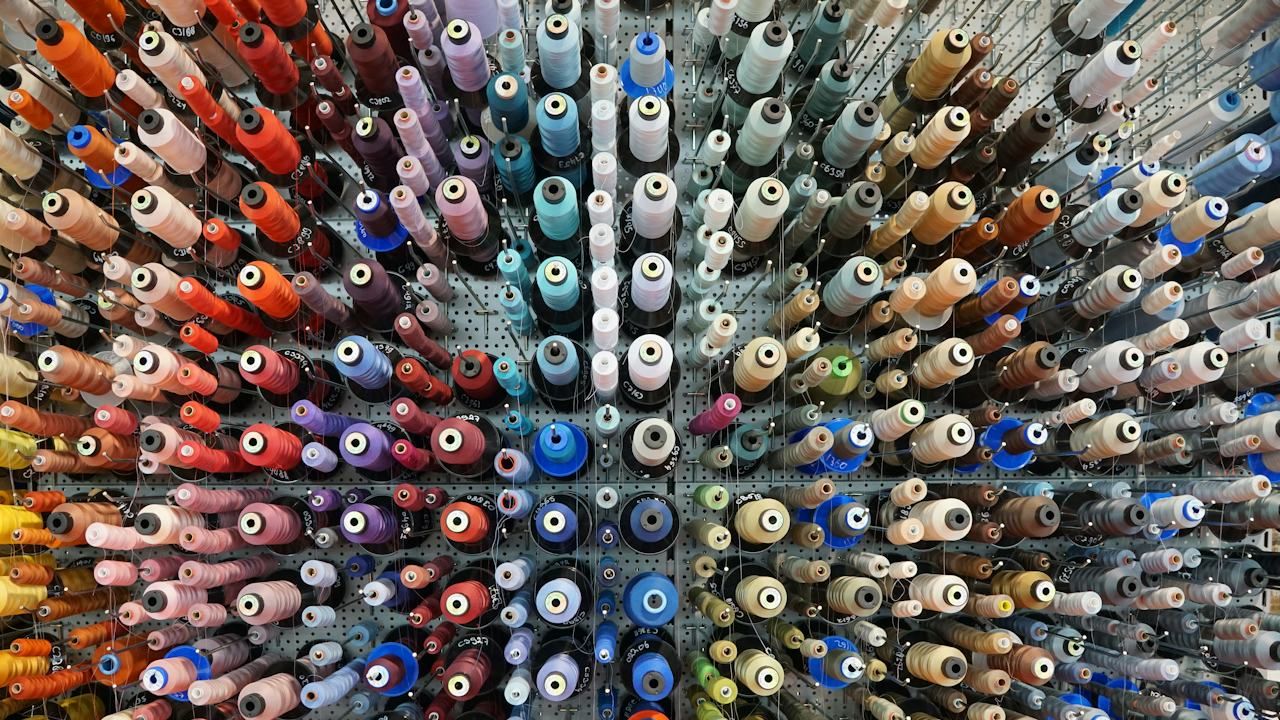 A pegboard wall seen head-on, filled with hundreds of sewing thread cones, roughly grouped by colour into blocks of orange and red, blue and white, beige and brown