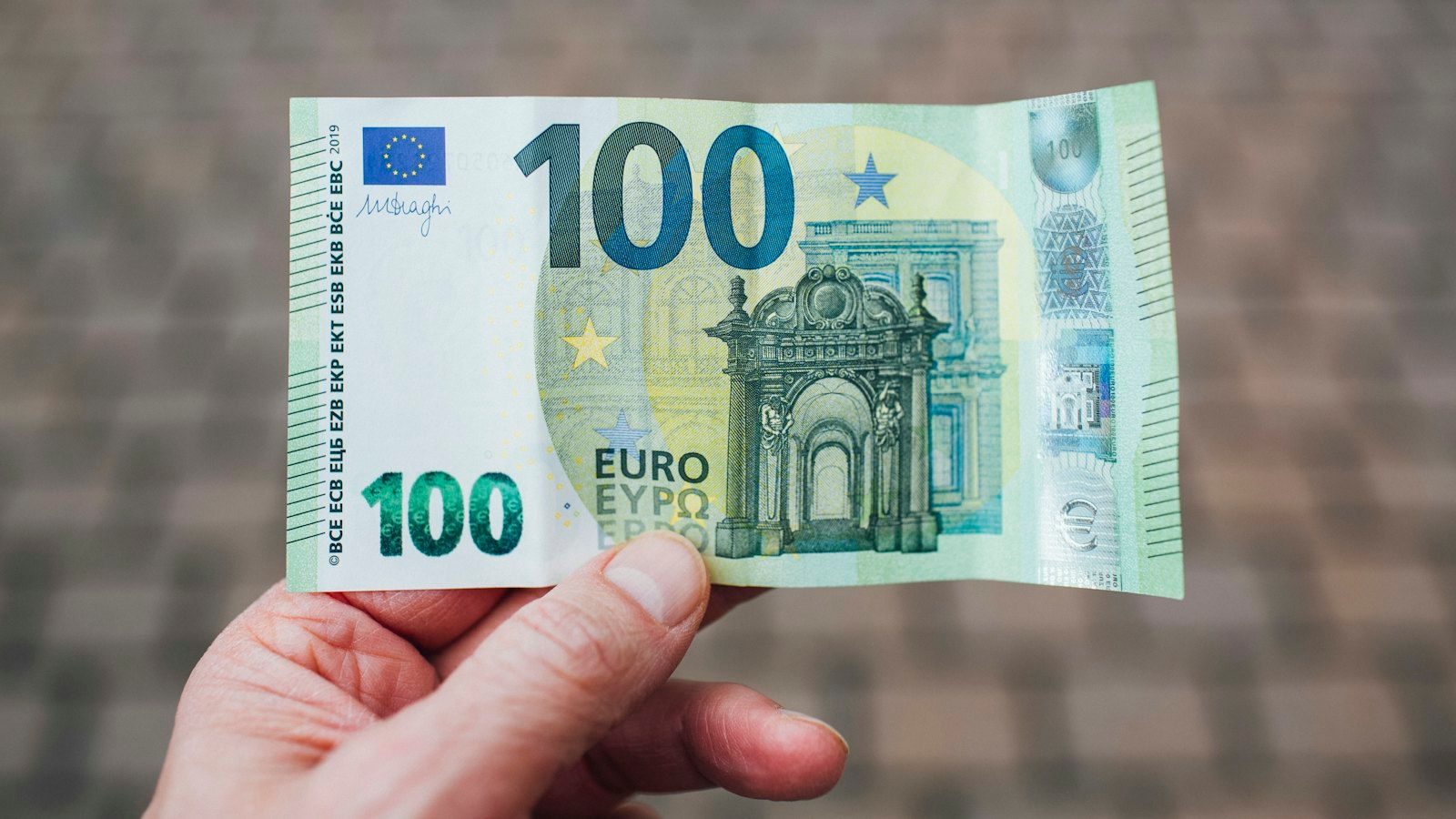 A hand holds a 100-euro banknote; on its right edge are the watermark area and a shiny security hologram, over a blurred grey-brown pavement