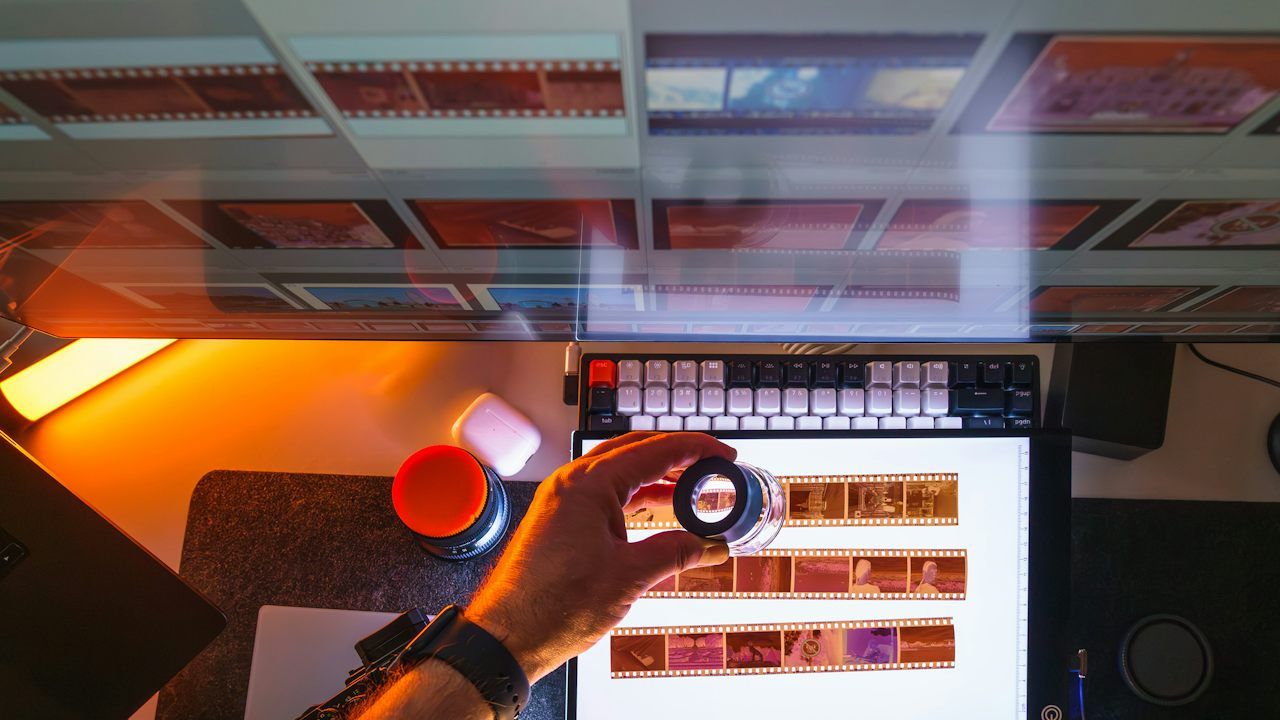Top-down view of a desk where a hand holds a loupe over colour film negatives on a lit light table, with rows of negatives in clear sleeves hanging above and an orange desk lamp on the left