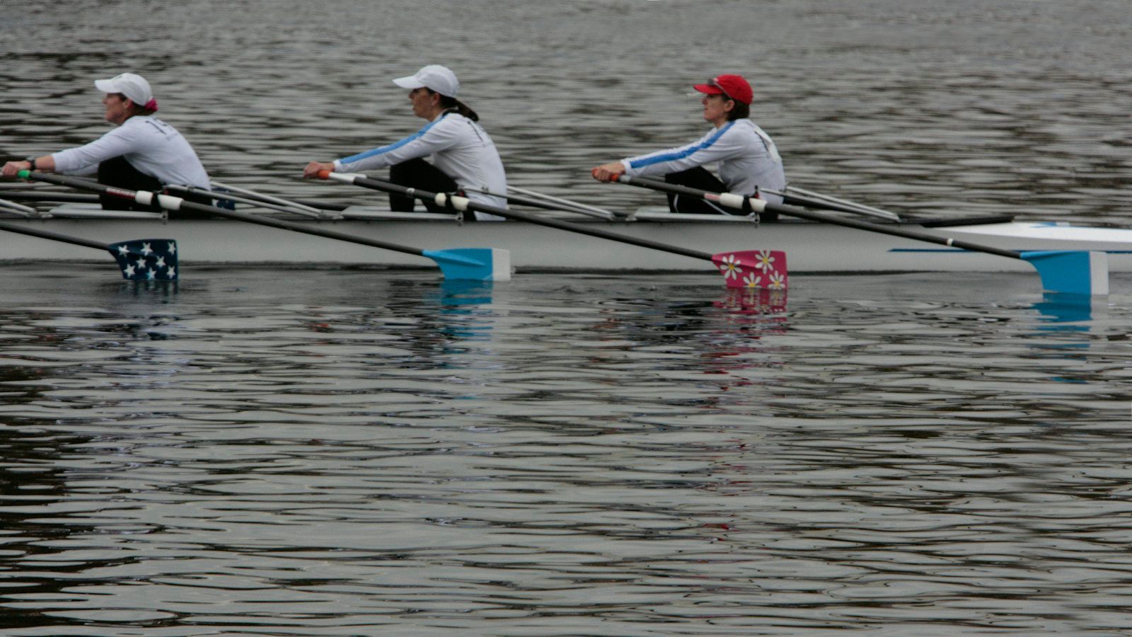 Three rowers in one boat lean forward together, their four differently coloured oar blades resting on the water at the same moment