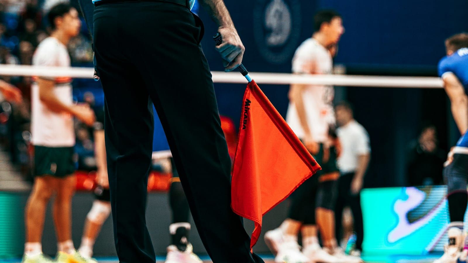 A volleyball line judge holding an orange-red flag at the edge of the court, players blurred behind