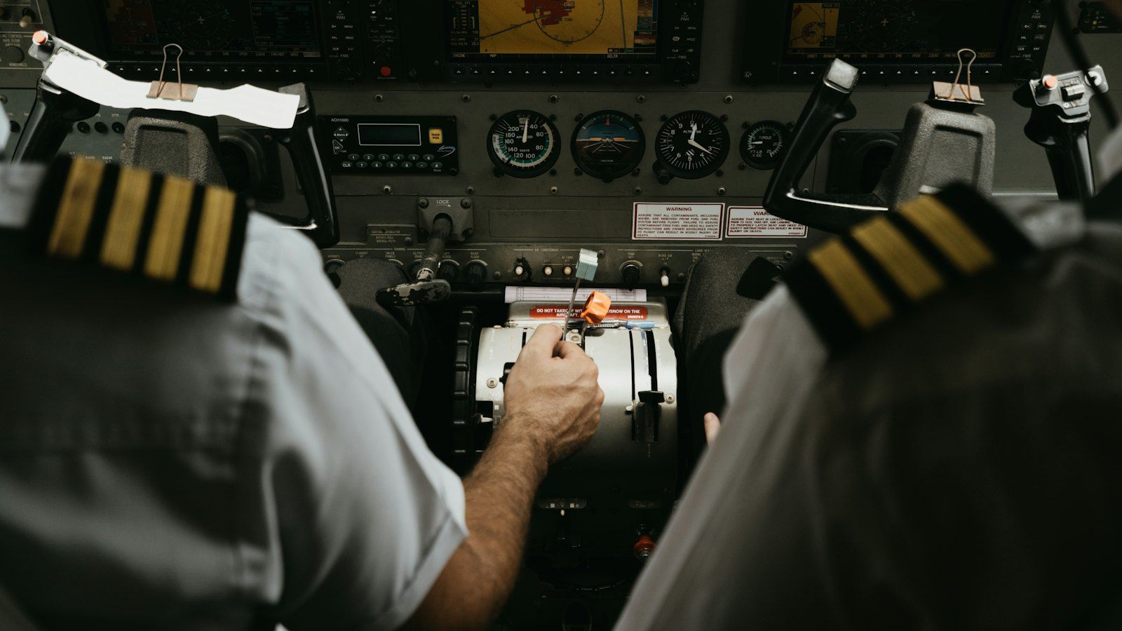 Over-the-shoulder view of a small aircraft cockpit, two pilots with four-stripe and three-stripe gold epaulettes, one hand on the throttle lever between them, a row of round gauges and two screens ahead