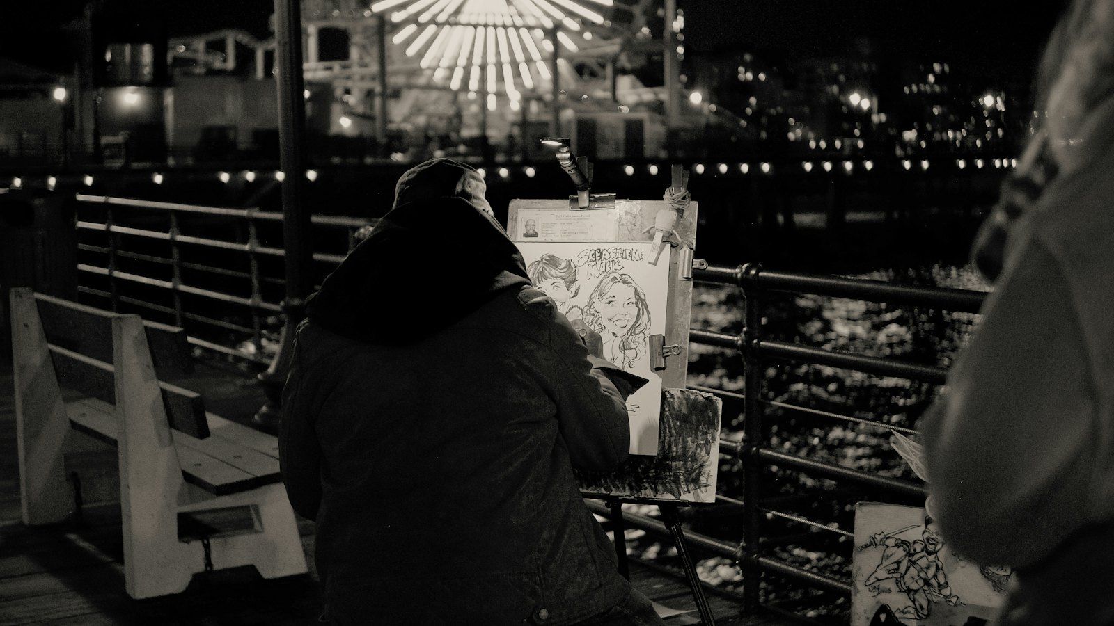 Black-and-white night shot of a pier, where a hooded street artist, back to the camera, draws a caricature on an easel with a lit Ferris wheel behind