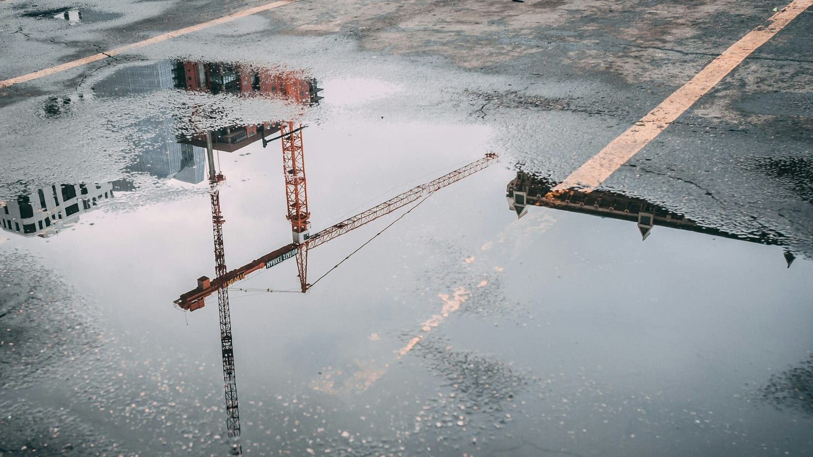 A puddle on wet asphalt reflects an orange tower crane and a few buildings upside down, next to yellow road markings