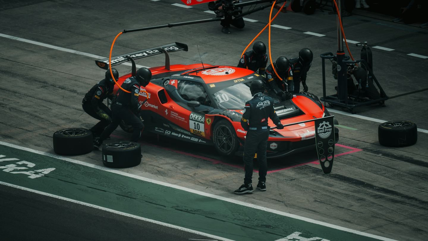 A red race car stopped in its pit box while five or six crew members in black suits and helmets change its tyres, with removed wheels on the ground