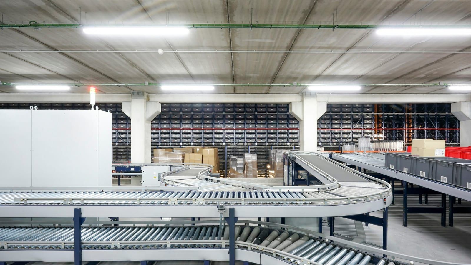 Inside a logistics sorting center, several silver roller conveyor lines curve and merge in the foreground, with a wall of racking and stacked cardboard boxes behind